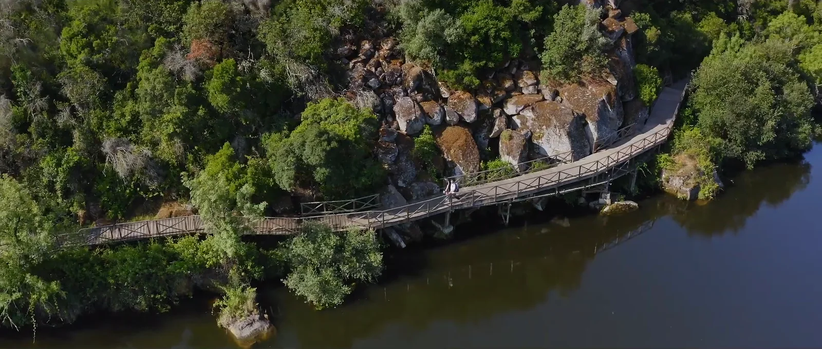 Duas pessoas num passadiço de madeira entre a encosta arborizada e o rio